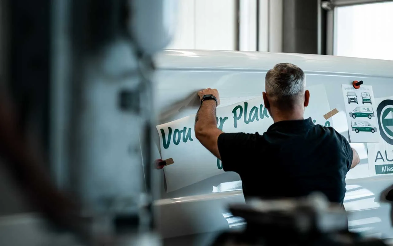Werbetechniker bringt in der Werkstatt eine grüne Folienbeschriftung auf einen weißen Transporter auf